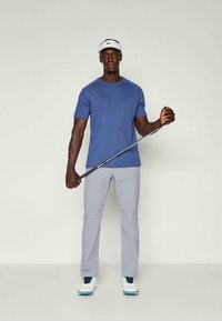 Atlet v modrém triku a světlých šedých kalhotách, držící golfovou hůl. Má na sobě bílé boty a bílou kšiltovku. Hladká látka, moderní střih.
