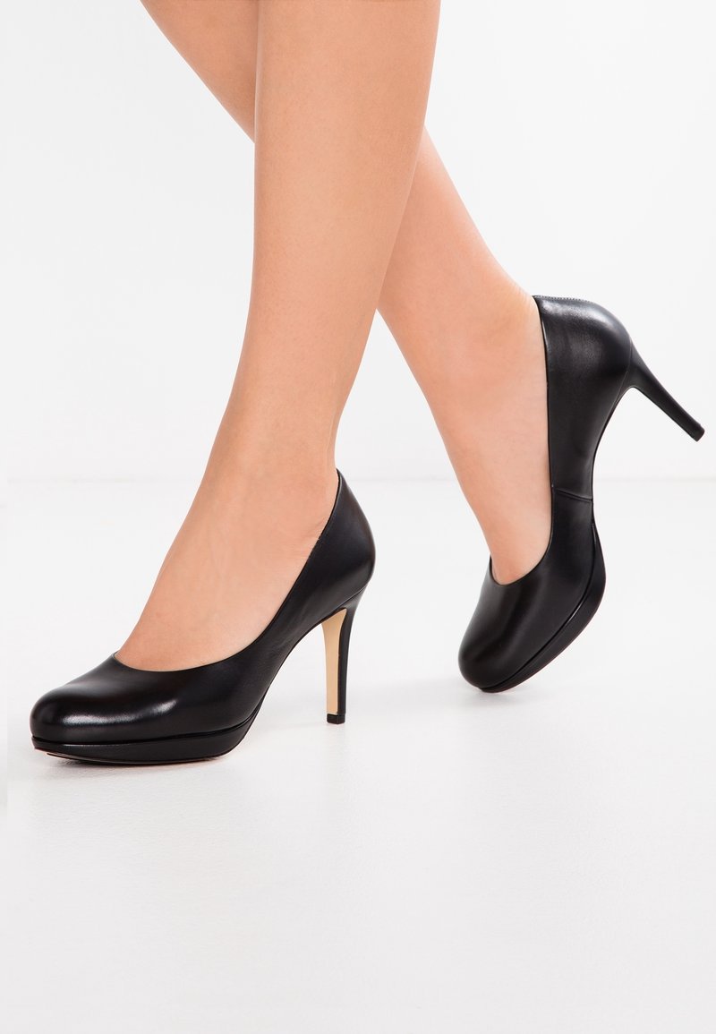 Jambes portant des escarpins en cuir noir à talons hauts, bout arrondi et talon fin, posées sur un fond blanc uni.