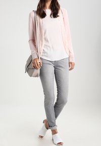 Ljusrosa kofta med spetskant, vit topp, grå jeans och vita öppna sandaler. Inkluderar en grå axelväska. Enkel design.