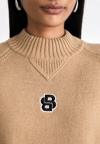 Suéter de malha bege com um alto colar canelado e um logótipo bordado em preto e branco no peito. Textura suave, design casual.