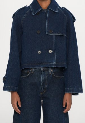 Personne portant une veste courte en denim bleu foncé à double boutonnage avec des manches larges et un jean taille haute assorti en denim.