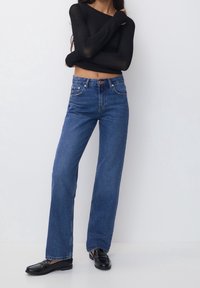 Pantalones vaqueros de mezclilla azul de pierna ancha con cintura alta, que cuentan con cinco bolsillos, costuras en contraste y cierre de cremallera. Combinados con mocasines negros.