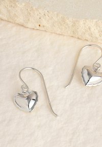 Srebrne kolczyki w kształcie serca z teksturowanym brzegiem i wypolerowaną powierzchnią, z hokerowym zapięciem do mocowania.