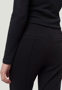 Pantalon noir ajusté en tissu lisse avec deux poches arrière et un détail de couture discret, associé à un haut noir à manches longues.