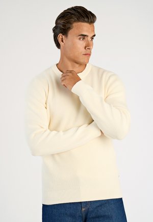 Maglione a coste di un crema chiaro con maniche lunghe e scollo rotondo, abbinato a jeans di denim blu scuro. La texture è liscia e aderente.