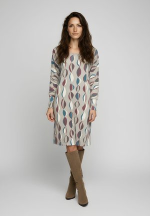 Mujer con cabello largo y oscuro, vestida con un vestido estampado hasta la rodilla y botas marrones hasta la rodilla, de pie frente a un fondo gris liso.