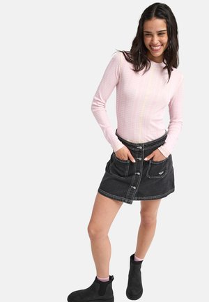 Jeune femme souriante, portant un haut à manches longues rose clair, une jupe en jean noire boutonnée et des bottines noires avec des chaussettes roses.