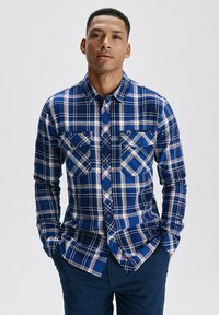 Niebieska koszula w kratę z guzikami, z czerwonymi, białymi i czarnymi akcentami; posiada dwie kieszenie na piersi i długie rękawy; wykonana z teksturowanej tkaniny.