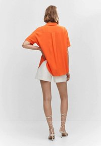 Pomarańczowa koszula z krótkim rękawem i plisą z tyłu, dopasowana do białych szortów. Modelka ma na sobie beżowe sandały na obcasie.