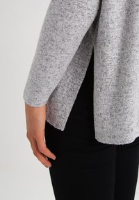 Maglione grigio a maglia con una finitura testurizzata, caratterizzato da maniche lunghe e un orlo spaccato. La mano di chi lo indossa è posizionata vicino al fianco.