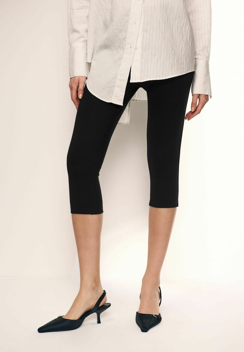 Leggings neri a capri realizzati con materiale elasticizzato. Design a vita alta con una texture liscia, abbinati a tacchi a punta slingback.