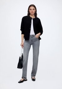 Femme portant un cardigan noir, un top blanc, un jean gris, des ballerines noires, tenant un sac à main noir avec une bandoulière chaîne, debout devant un fond uni.