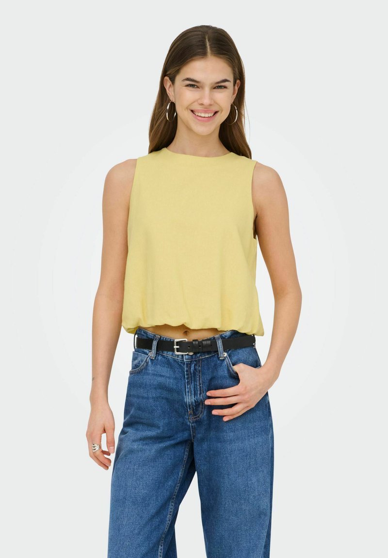 Jeune femme souriante, portant un haut jaune sans manches légèrement relevé au-dessus du nombril, un jean bleu taille haute avec une ceinture noire, et des boucles d'oreilles créoles.