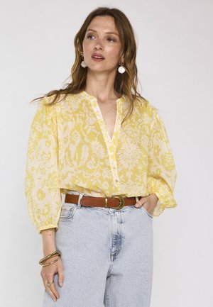 Femme aux longs cheveux bruns portant un chemisier jaune à fleurs avec des manches bouffantes, un jean bleu clair, une ceinture marron, des boucles d'oreilles anneaux et des bracelets.