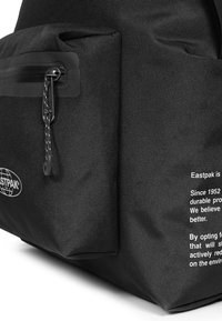 Černý batoh Eastpak s rohem, přední kapsou na zip, reflexní úchytkou na zip a bílým potiskem značky a zprávou na textilu.