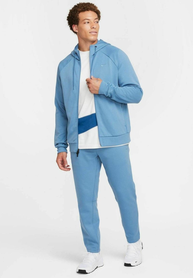 Helles blaues Sportanzug aus weichem Material, bestehend aus einer Kapuzenjacke mit Reißverschluss, einem weißen Shirt mit blauen Akzenten und passender Hose. Weiße Sneakers.