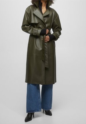 Personne portant un long trench-coat en cuir vert foncé, un jean bleu à jambes larges et des bottines noires à talons hauts sur un fond uni.