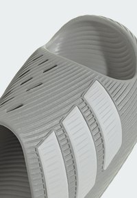 Primer plano de una sandalia gris sin cordones con superficie texturizada y tres rayas blancas inclinadas en la parte superior.