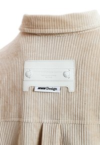 Tela de pana beige con estrías verticales, que presenta una etiqueta de cuero blanco adjunta en el cuello con detalles en texto negro.