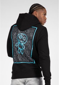 Sudadera negra con un gran gráfico turquesa en la espalda, que presenta un personaje de dibujo animado y un patrón de texto repetitivo. Tejido suave, ajuste casual.