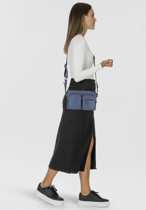 Bolso bandolera de cuero azul con dos bolsillos frontales y un compartimento con cremallera. Cuenta con una correa ajustable. Se lleva con una falda negra y zapatos.