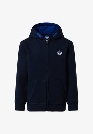 Garçon en sweat à capuche bleu marine, t-shirt bleu, shorts bleu marine, baskets blanches, chaussettes blanches, tenant une casquette de baseball bleu marine sur un fond uni.