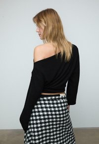 Crni skraćeni pulover s dizajnom bez ramena, dugim rukavima. Uparen s suknjom u crno-bijeli karirani uzorak. Glatka tekstura, ležerni stil.