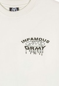 Jasno beżowa koszulka bawełniana z okrągłym dekoltem, z czarnym logo w formie cieknącego napisu "INFAMOUS GRMY" umieszczonym na środku z przodu.