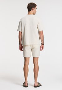 Beige cotton t-shirt and shorts set, relaxed fit. T-shirt has short sleeves and crew neck; shorts are plain. Worn with brown sandals.