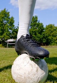 Svart fotballsko med teksturert design og grønne detaljer hviler på en slitt hvit fotball på gress, med trær og et mål i bakgrunnen.