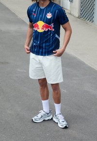 Maglia sportiva a righe blu con accenti rossi e logo, abbinata a pantaloncini bianchi e sneakers argento massicce. In piedi su una superficie di cemento.