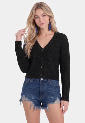 Young woman with long blonde hair wearing blue tassel earrings, a black buttoned ribbed sweater, and distressed denim shorts.