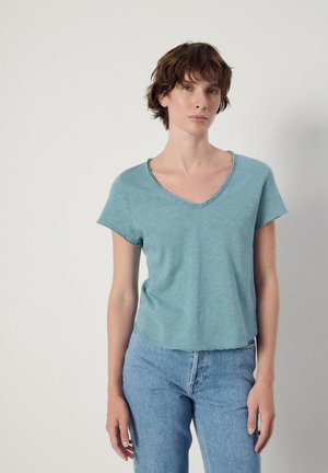 Jong persoon met kort bruin haar draagt een lichtblauw T-shirt met korte mouwen en V-hals en een hoge taille blauwe jeans, staand tegen een effen muur.