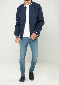 Marine bomberjack met een ritssluiting aan de voorkant, witte gestreepte manchetten, gecombineerd met lichtblauwe jeans en donkere sneakers. Gladde stof, casual ontwerp.