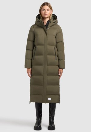 Olijfgroene pufferjas met een lange uitstraling, horizontale stiksels, ritszakken aan de zijkant en een capuchon, gedragen met zwarte kniehoge laarzen.