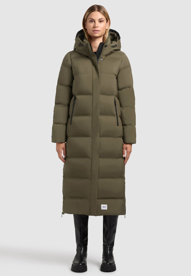 Olijfgroene pufferjas met een lange uitstraling, horizontale stiksels, ritszakken aan de zijkant en een capuchon, gedragen met zwarte kniehoge laarzen.