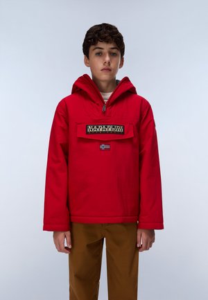 Teenagedreng iført en rød hættetrøje med en frontlomme og logo, brune bukser, stående mod en ensartet lys baggrund.
