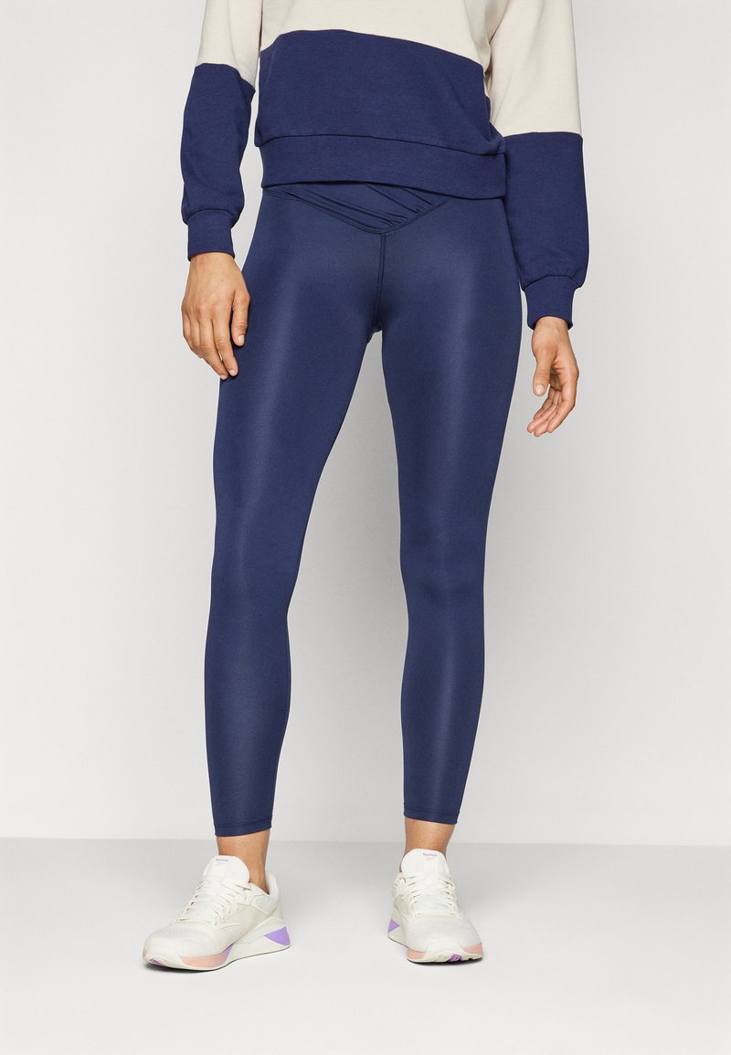 Granatowe legginsy wykonane z elastycznej tkaniny o błyszczącym wykończeniu. Posiadają wysoki pas i gładką teksturę. W połączeniu z jasnymi trampkami.