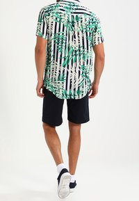 Kurzarm-Hemd mit Kragen, bedruckt mit grünem tropischem Blattmuster und marineblauen vertikalen Streifen. Getragen mit marineblauen Shorts und weißen Socken.