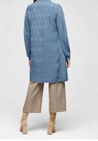 Lungo cappotto lavorato a maglia blu con una texture ondulata, design con colletto e spacchi laterali, indossato sopra pantaloni a vita alta con fantasia e stivali beige fino al ginocchio.
