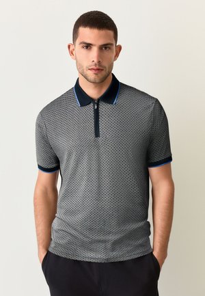 Polo a maniche corte color grigio scuro con motivo a puntini e colletto blu navy. Presenta una scollatura con zip e dettagli blu sulle maniche.