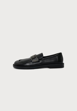 Mocassino in pelle nera con tacco basso e cuciture decorative sulla fascia superiore, mostrato di profilo su sfondo uniforme.