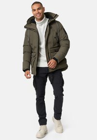 Doudoune vert olive avec capuche, poches avant et cordons de serrage ajustables, portée sur un pull en tricot blanc et un pantalon cargo noir.