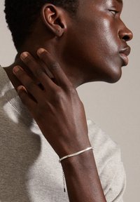 Silver bracelet with a sleek design, featuring a delicate chain and a textured surface, worn on a dark-toned wrist against gray fabric.