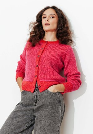 Femme aux cheveux bouclés portant un cardigan rouge vif boutonné et un jean gris taille haute, debout les mains dans les poches devant un fond blanc.