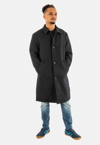 Abrigo de lana negro con cuello, botones frontales y bolsillos laterales. Lleva puesto sobre una blusa negra y jeans de mezclilla azul claro, con zapatillas azul y marrón.