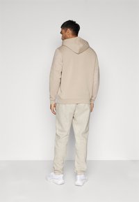 Beige hoodie met lichtere beige mouwen, gecombineerd met lichtgrijze sweatpants. De outfit wordt aangevuld met witte sportschoenen.