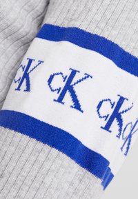 Manica di maglione grigio a maglia con una banda bianca con il testo blu "ck" in ripetizione, sfondo texturizzato con motivo a coste.