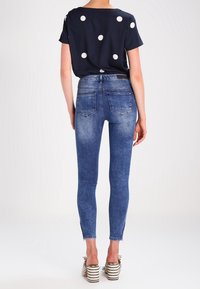 Top de lunares azul marino con mangas cortas, combinado con vaqueros ajustados azules. Los vaqueros tienen una textura desgastada y bolsillos traseros. Sandalias de tacón alto a rayas.