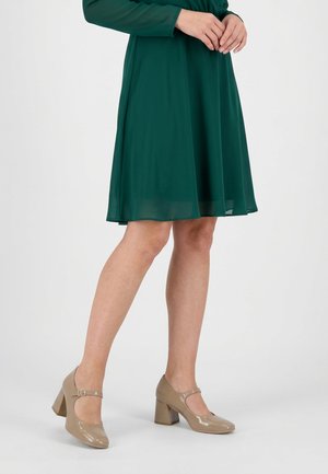 Žena stojící se zkříženýma nohama, oblečená v tmavě zelených šatech délky ke kolenům a béžových lakovaných botách Mary Jane na blokovém podpatku.